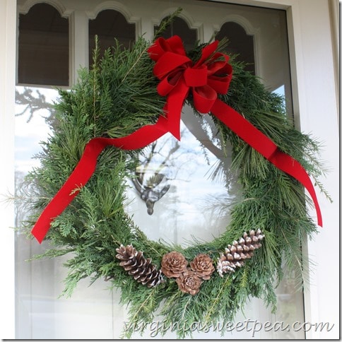 Live greenery wreath made with a variety of pine species and decorated with red ribbon and pine cones.