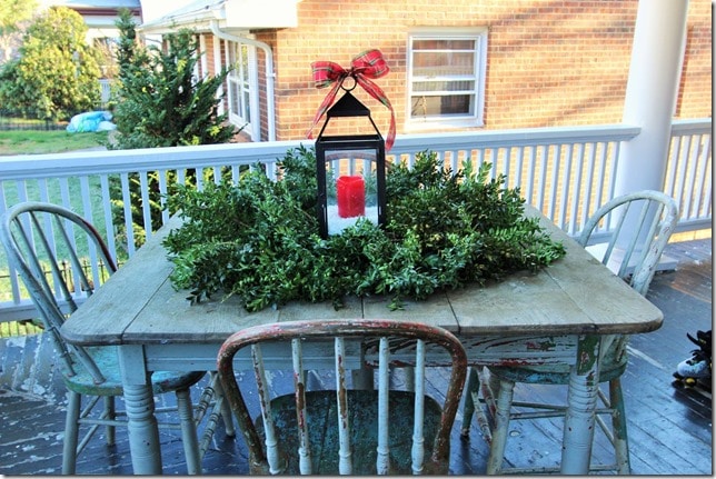 A chippy table and chairs on a porch looks festive for Christmas with a wreath and lantern.