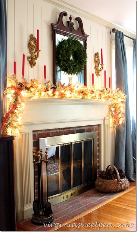 Christmas mantel with a gold lighted garland, red ribbon and candles, and snow covered pine cones. Vintage scones and an antique mirror with a Christmas wreath make this area look so inviting for Christmas.