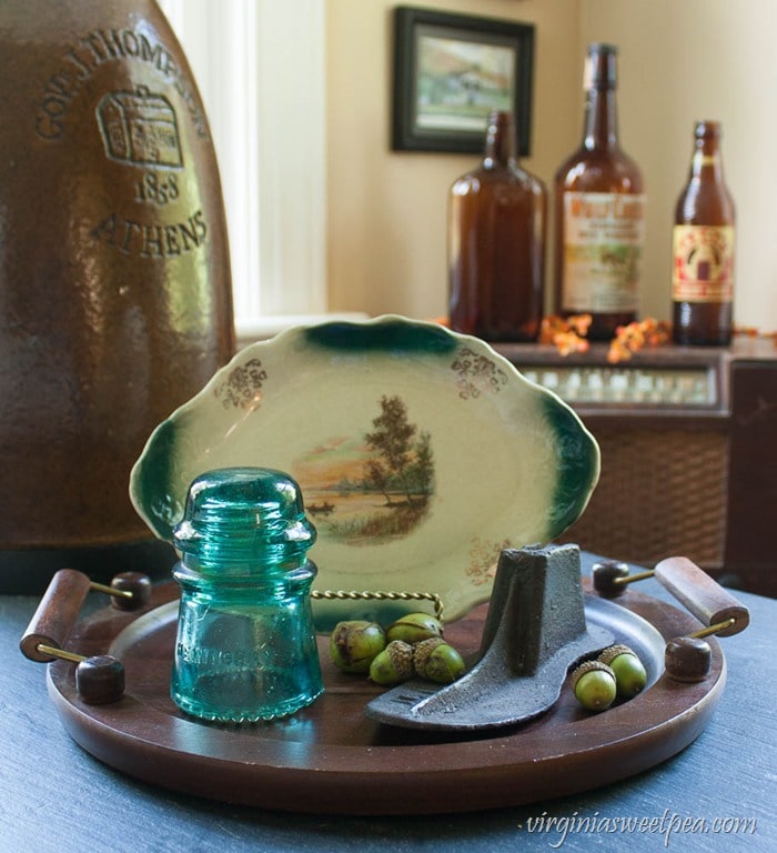 Fall decor with a wooden tray decorated with a small platter with a fall scene, an antique cast iron shoe form, a glass insulator, and in the background three vintage brown bottles.