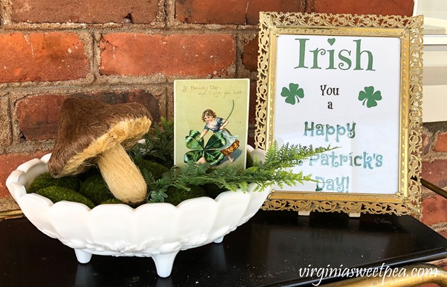 St. Patrick's Day decor with a milk glass composte filled with a mushroom, faux ferns, and a vintage St. Patrick's Day card displayed beside a framed St. Patrick's Day printable