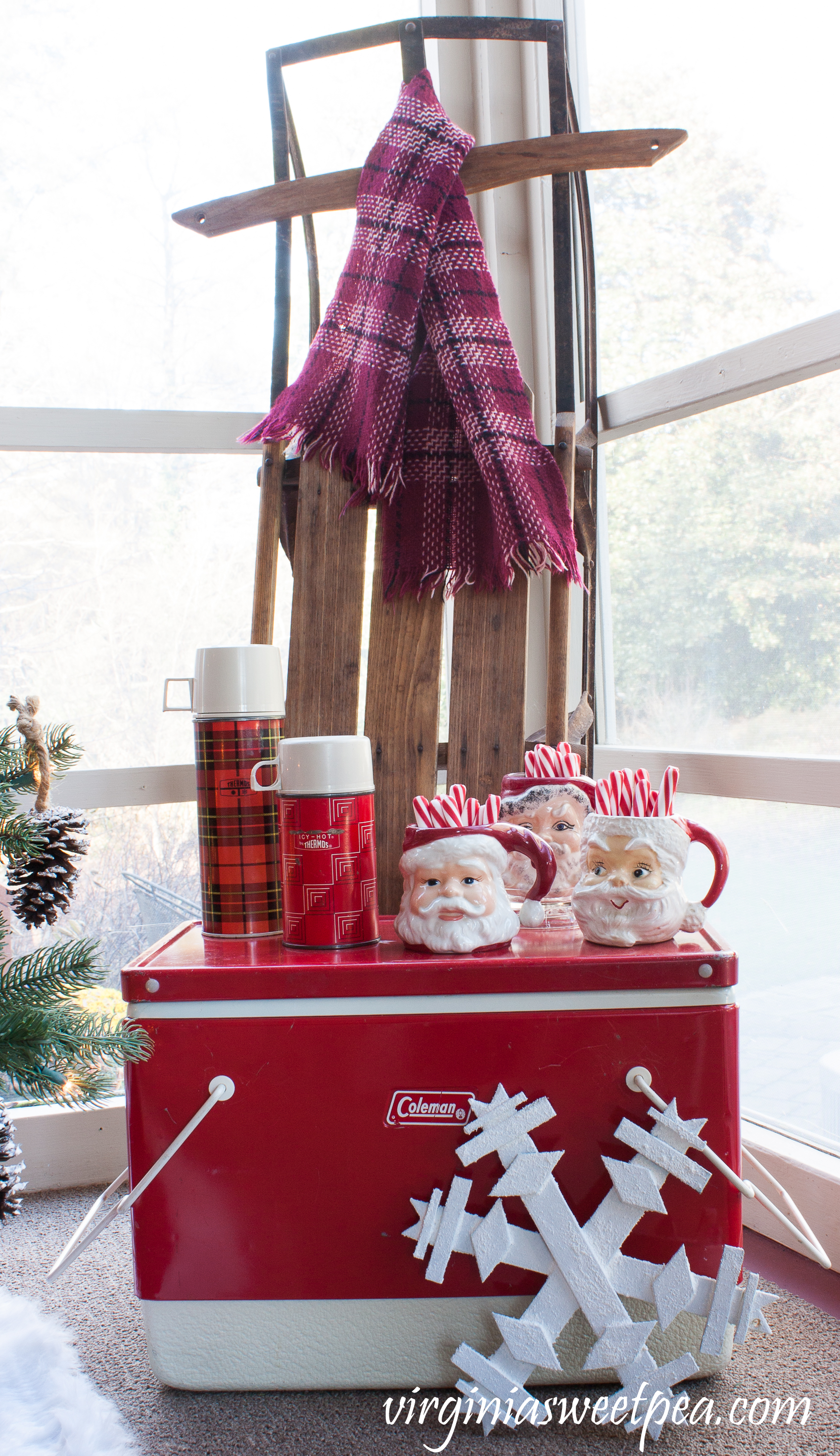 Christmas Porch Vignette - A vintage Coleman cooler is topped with vintage thermoses and Santa mugs. A vintage sled is adorned with a vintage scarf. #christmas #christmasdecorations #christmasporch #vintage #vintagechristmas