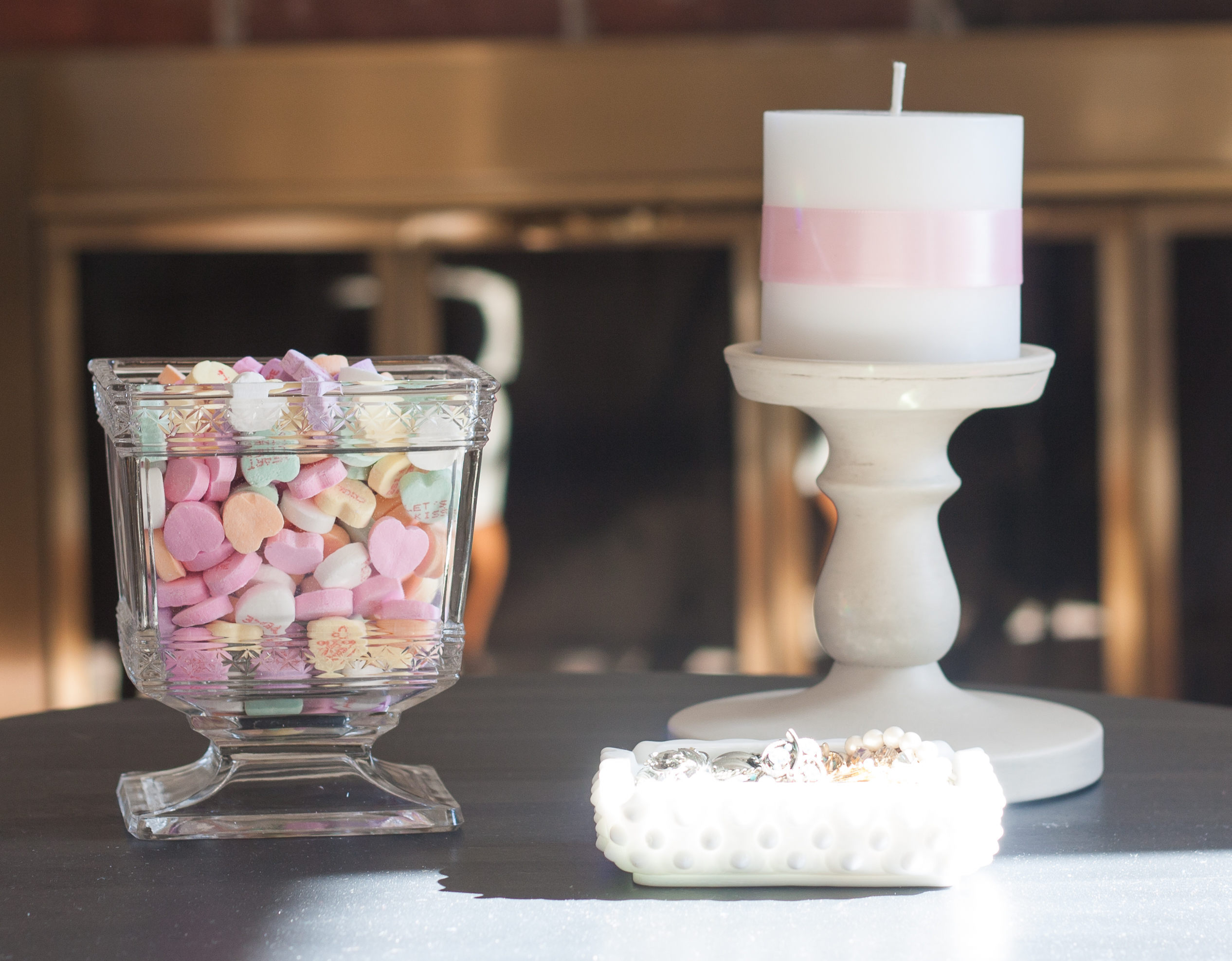 Valentine's Day Vignette with Conversation Hearts in an antique glass vase; vintage costume jewelry in a milk glass ashtray; and a candle made romantic with the addition of pink ribbon. #valentinesday #valentinesdayvignette #valentinesdaydecor #conversationhearts #milkglass