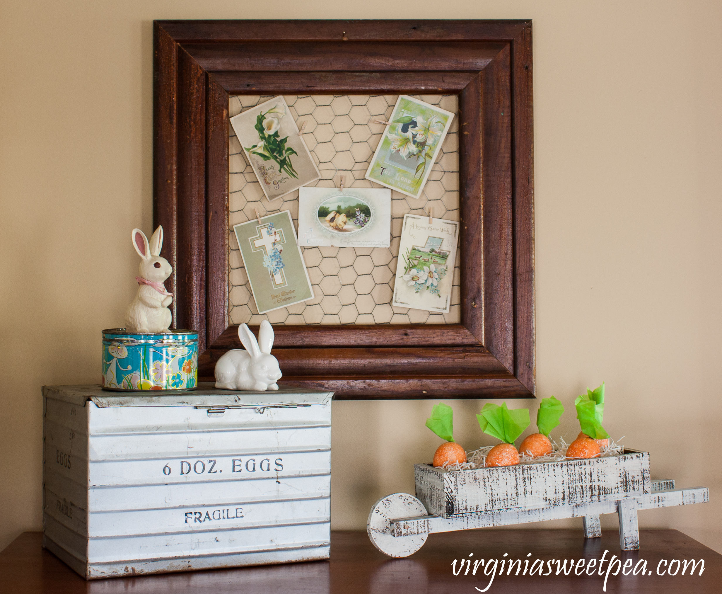 Easter Display with a vintage egg crate, 1960's candy tin, bunnies, early 1900's Easter postcards, and a wheelbarrow filled with eggs that look like carrots.