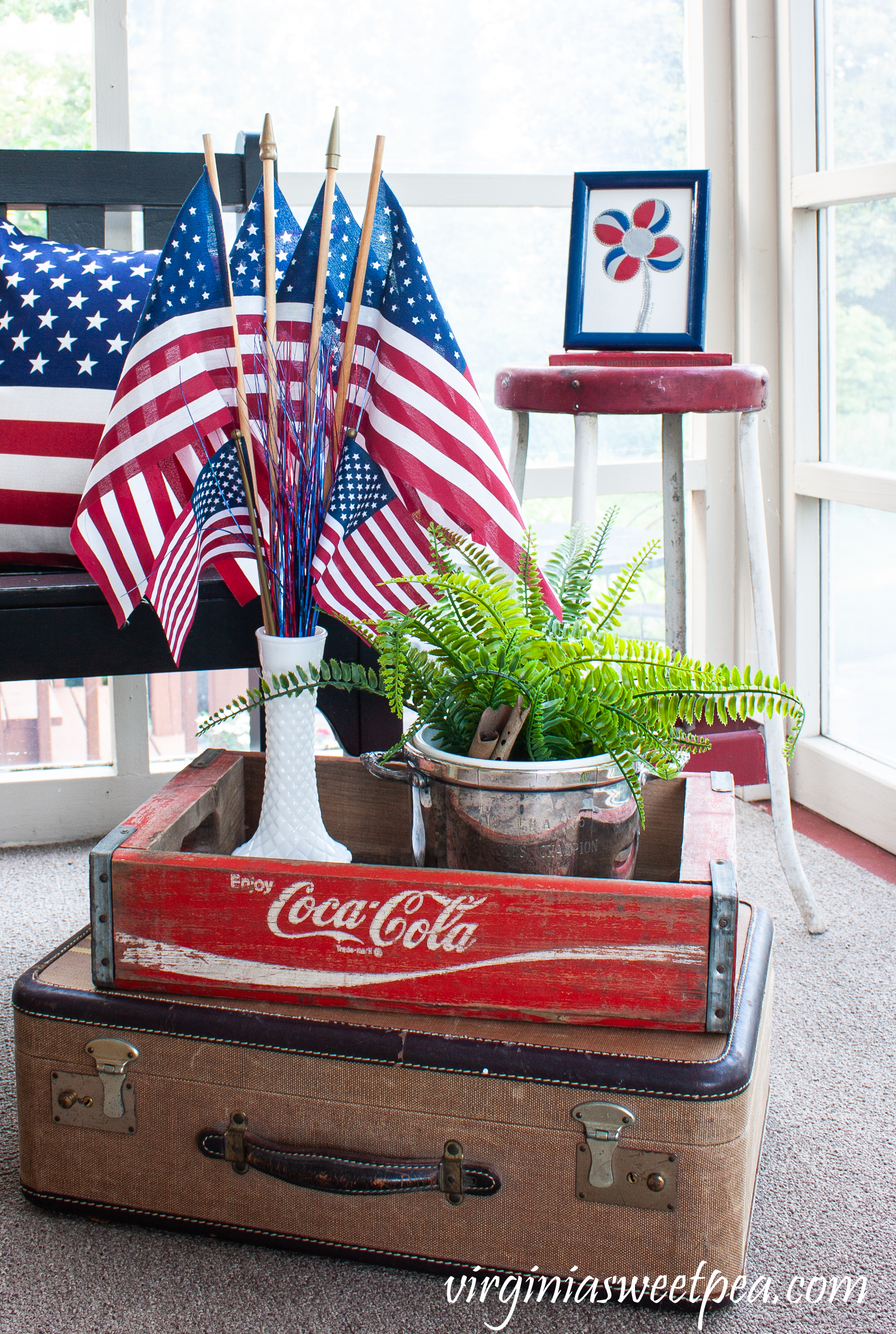 Patriotic display with a Coke crate, vintage suitcase, and milk glass vase filled with flags.