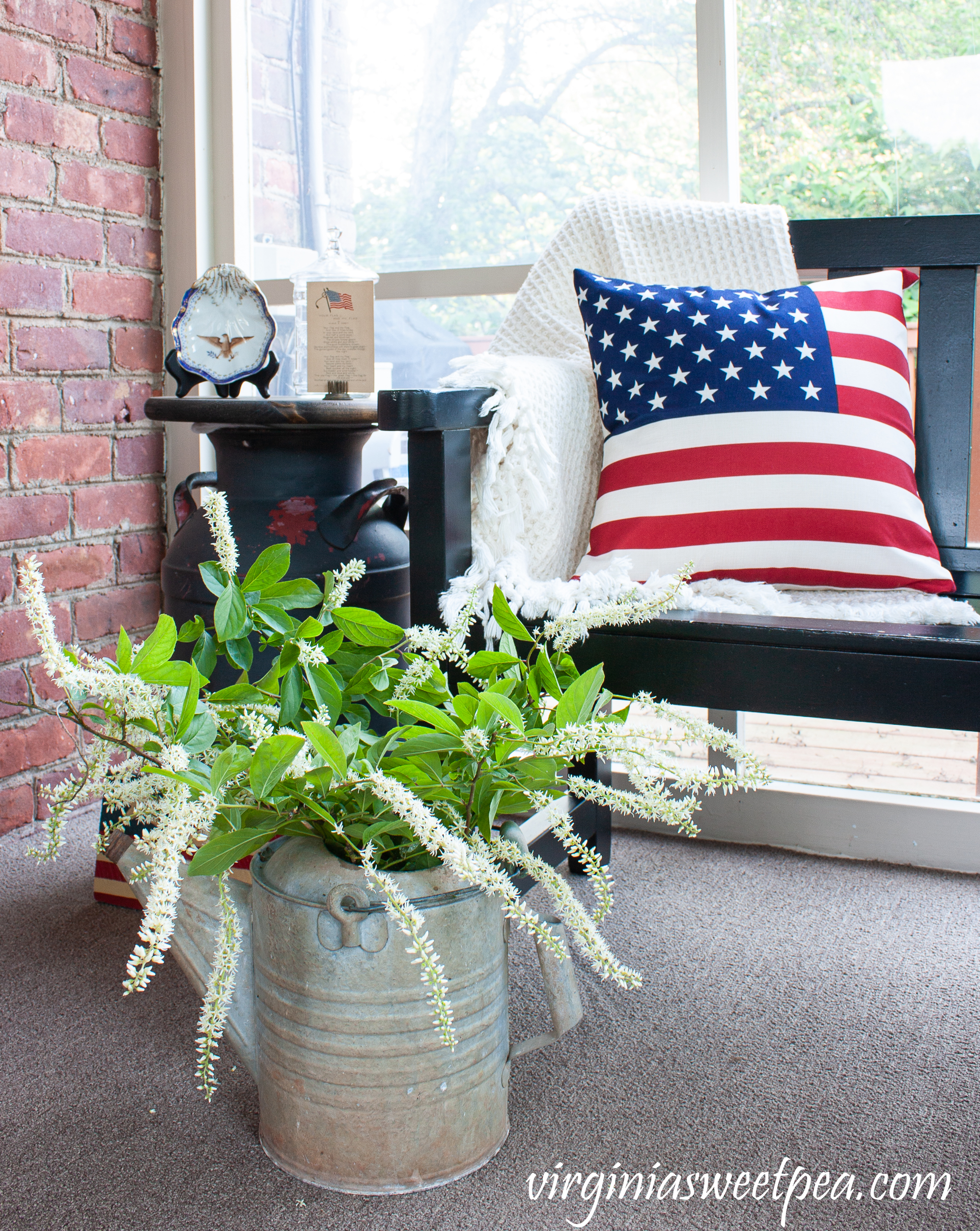 Patriotic porch decor with an American flag pillow and a vintage watering can filled with Virginia Sweetspire.