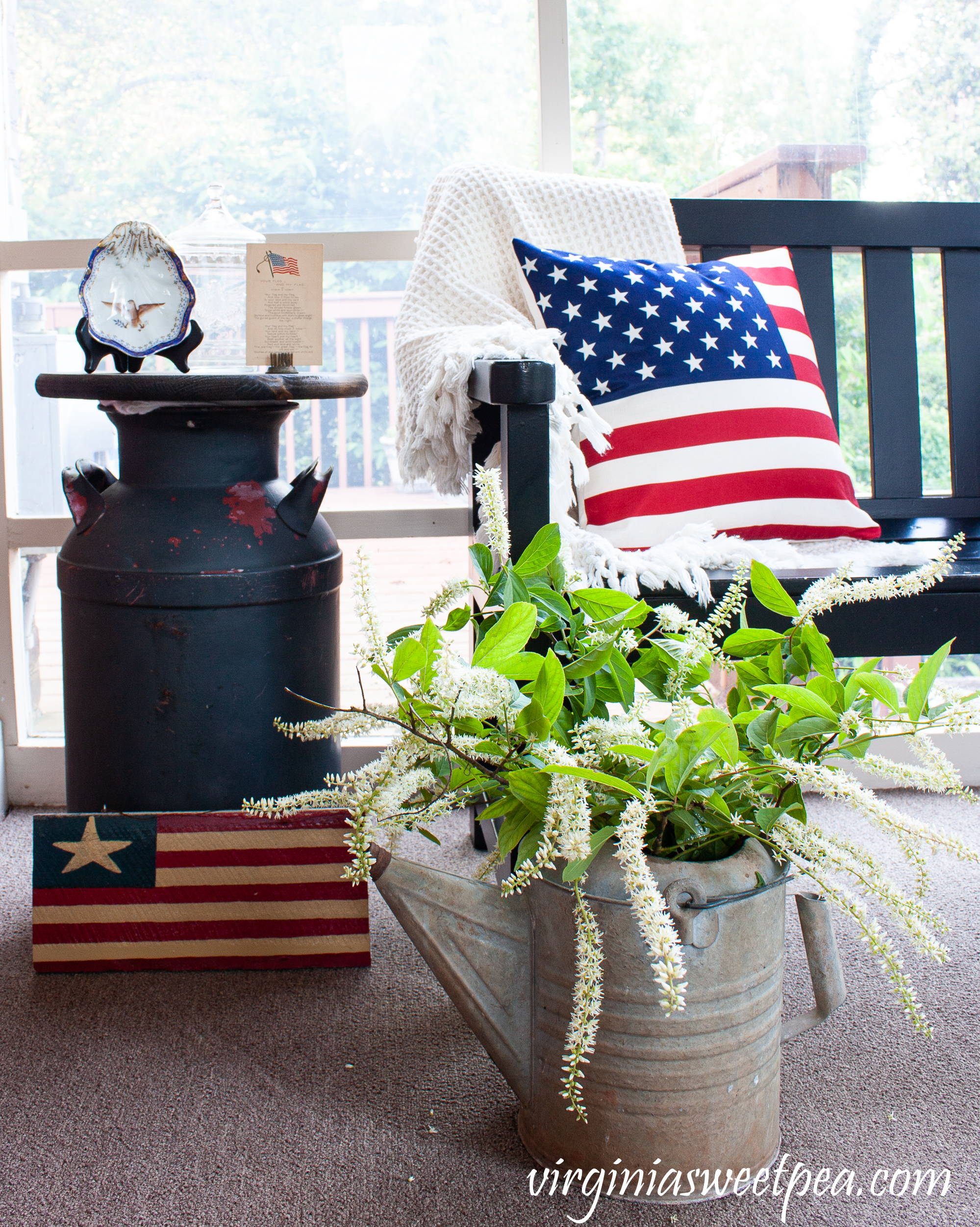 Patriotic porch decor including American flag pillows, a flag sign, a vintage milk can made into a table, and a vintage watering can.