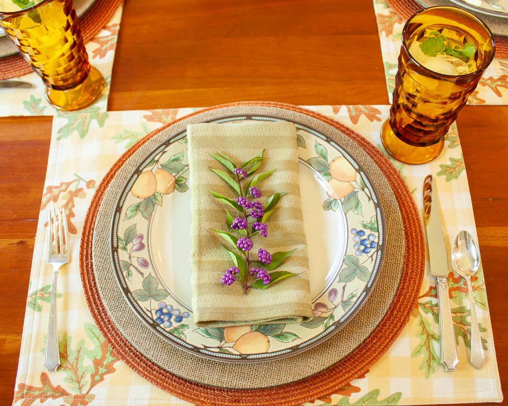 Table setting for fall with placemats printed with leaves, a round orange placemat, burlap charger, Mikasa Garden Harvest dishes, vintage silver, amber Anchor Hocking vintage glasses, and a napkin with a sprig of Beautyberry on top.