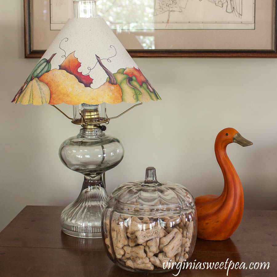 Antique lamp with a fall themed lamp shade, clay gourd shaped to look like a goose and a clear pumpkin lidded jar filled with Milk Bones for the dog.