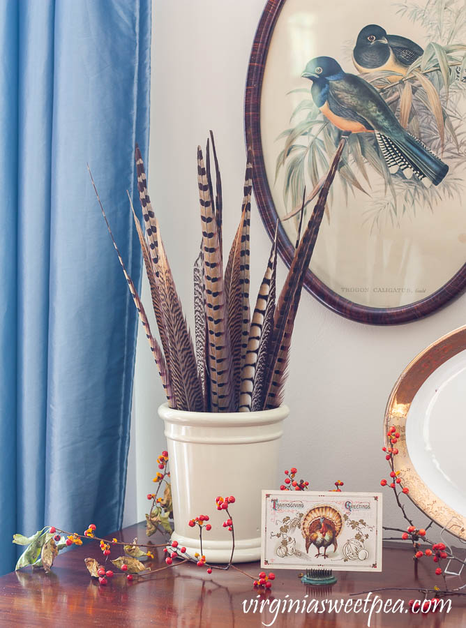 Thanksgiving vignette with a crock filled with pheasant feathers, a Thanksgiving postcard, and Bittersweet.