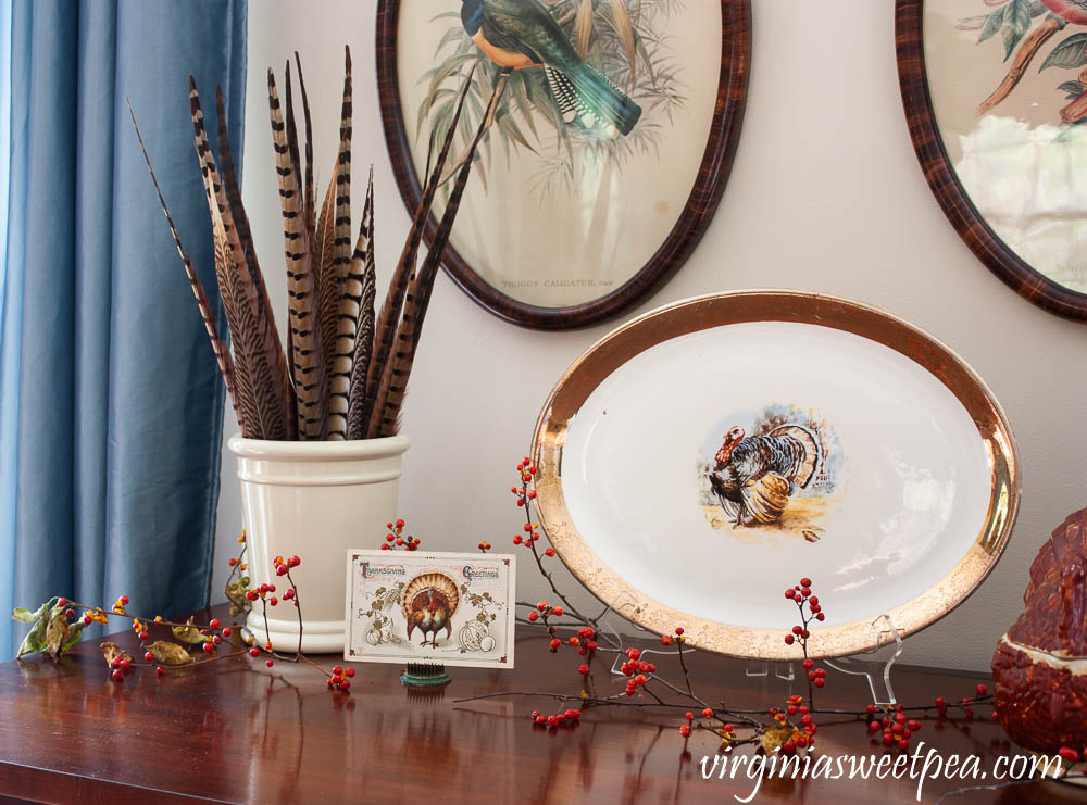 Thanksgiving vignette with a crock filled with pheasant feathers, a Thanksgiving postcard, and a vintage turkey platter.