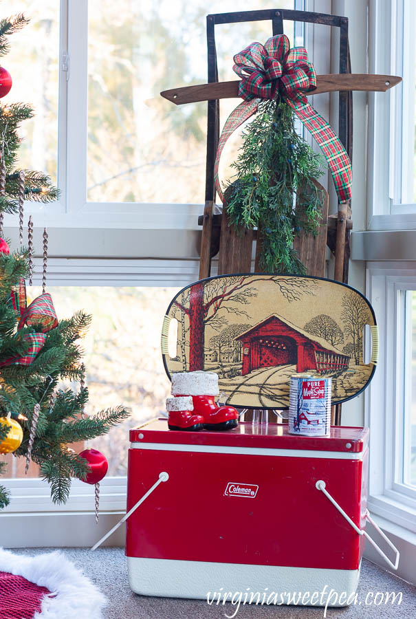 A Very Vintage Christmas on the Porch - Vintage sled decorated with a bow and greenery, vintage Coleman cooler with vintage Santa boots, a vintage covered bridge tray, and a maple syrup scented candle in a maple syrup can