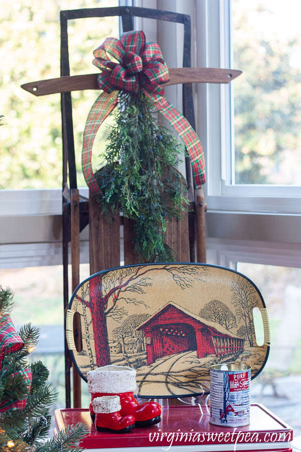 A Very Vintage Christmas on the Porch - Vintage sled decorated with a bow and greenery, vintage Coleman cooler with vintage Santa boots, a vintage covered bridge tray, and a maple syrup scented candle in a maple syrup can