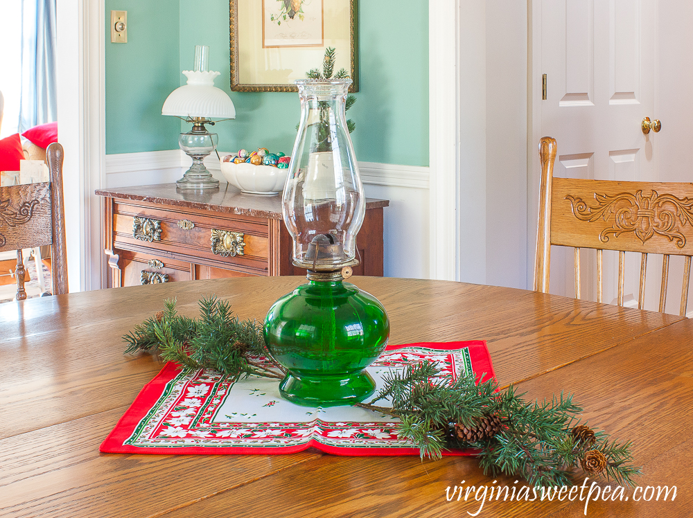 A Very Vintage Christmas in the Dining Room - Oil lamp with a green base used as a table centerpiece with a vintage Christmas napkin and greenery.