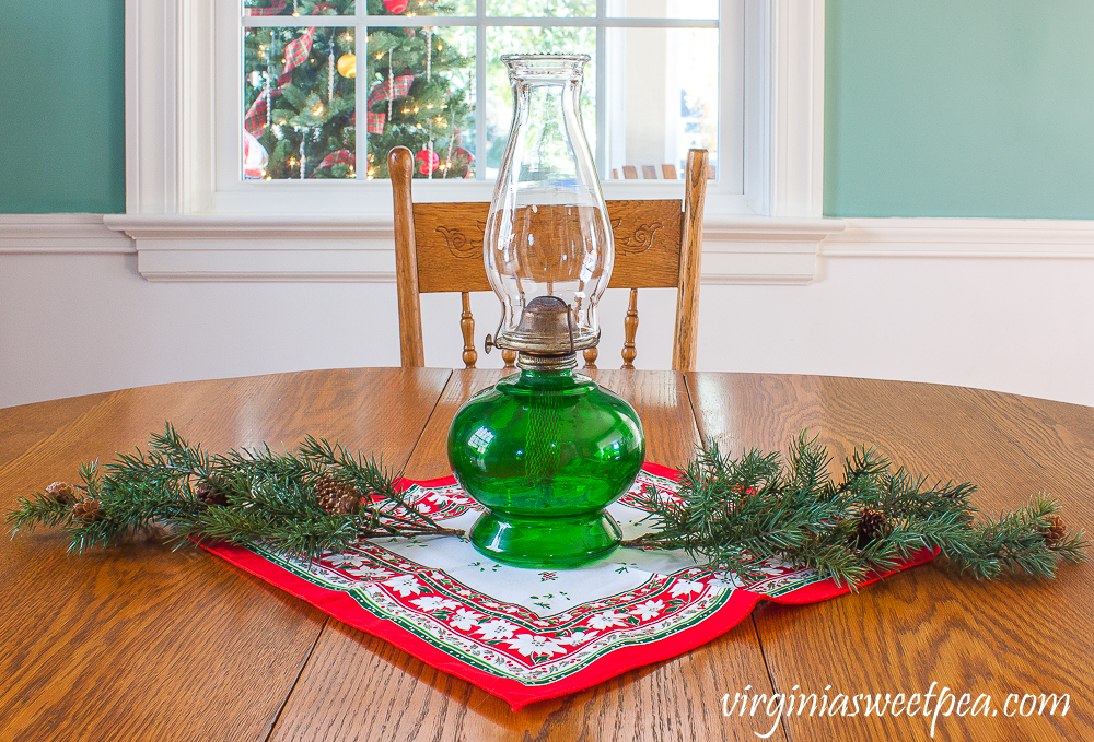 A Very Vintage Christmas in the Dining Room - Oil lamp with a green base used as a table centerpiece with a vintage Christmas napkin and greenery.