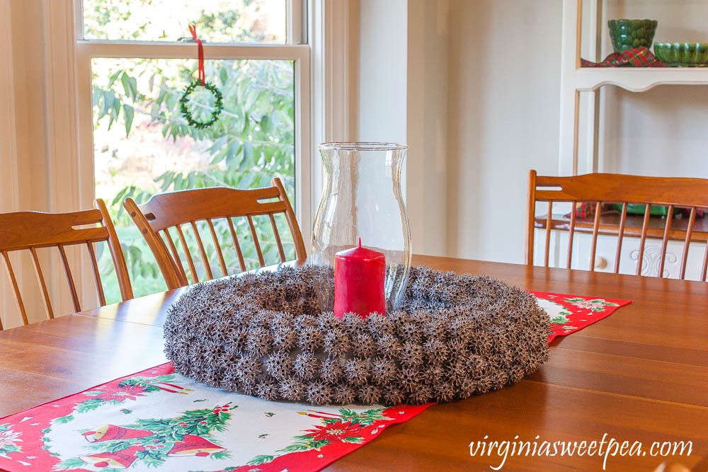 Wreath made from Sweet Gum balls with a glass hurricane and red candle on top of a vintage table runner.