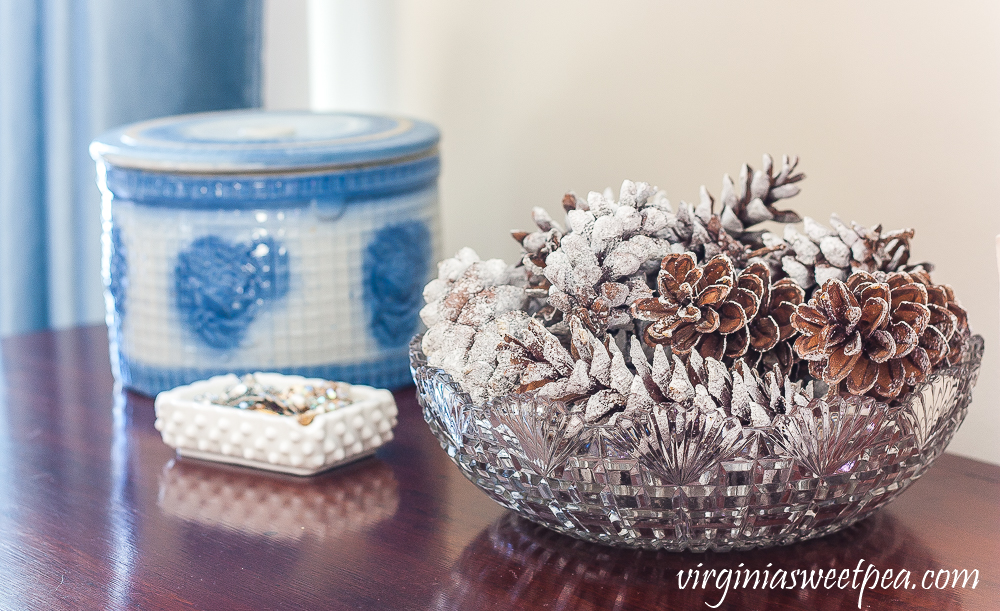 Blue and white antique crock with lid, milk glass ashtray filled with vintage jewelry, antique cut glass bowl filled with pine cones coated in faux snow.