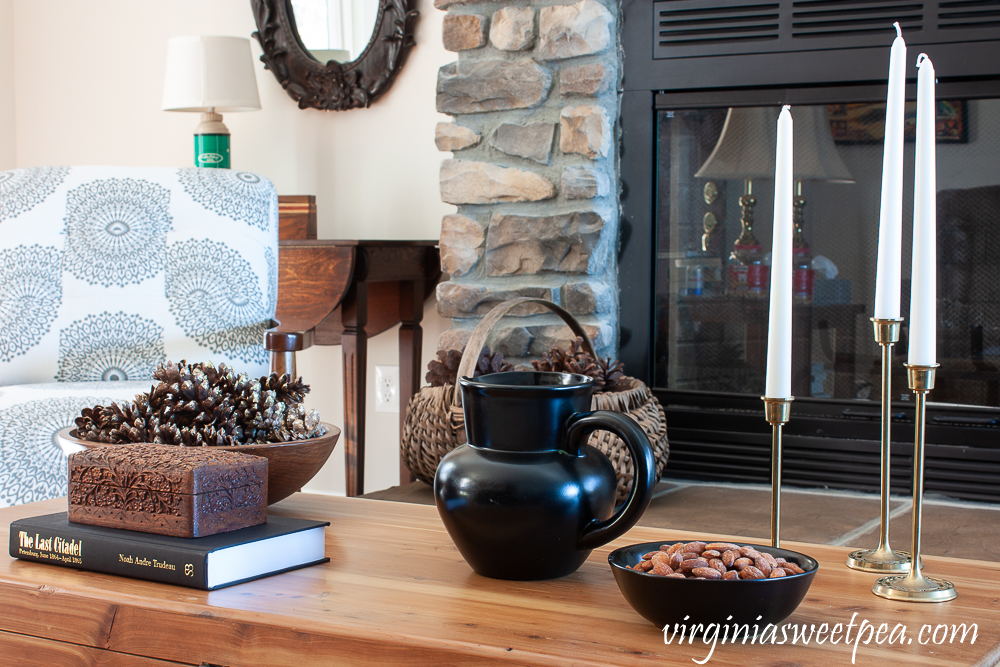 Winter coffee table decor with wood bowl filled with pine cones, carved wood box, black pitcher, black bowl with almonds, and three brass candlesticks with white candles