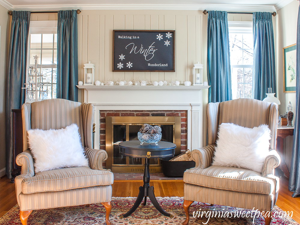 Winter mantel, wing back chairs with white fur pillows, a table with an antique glass compote filled with silver pinecones.