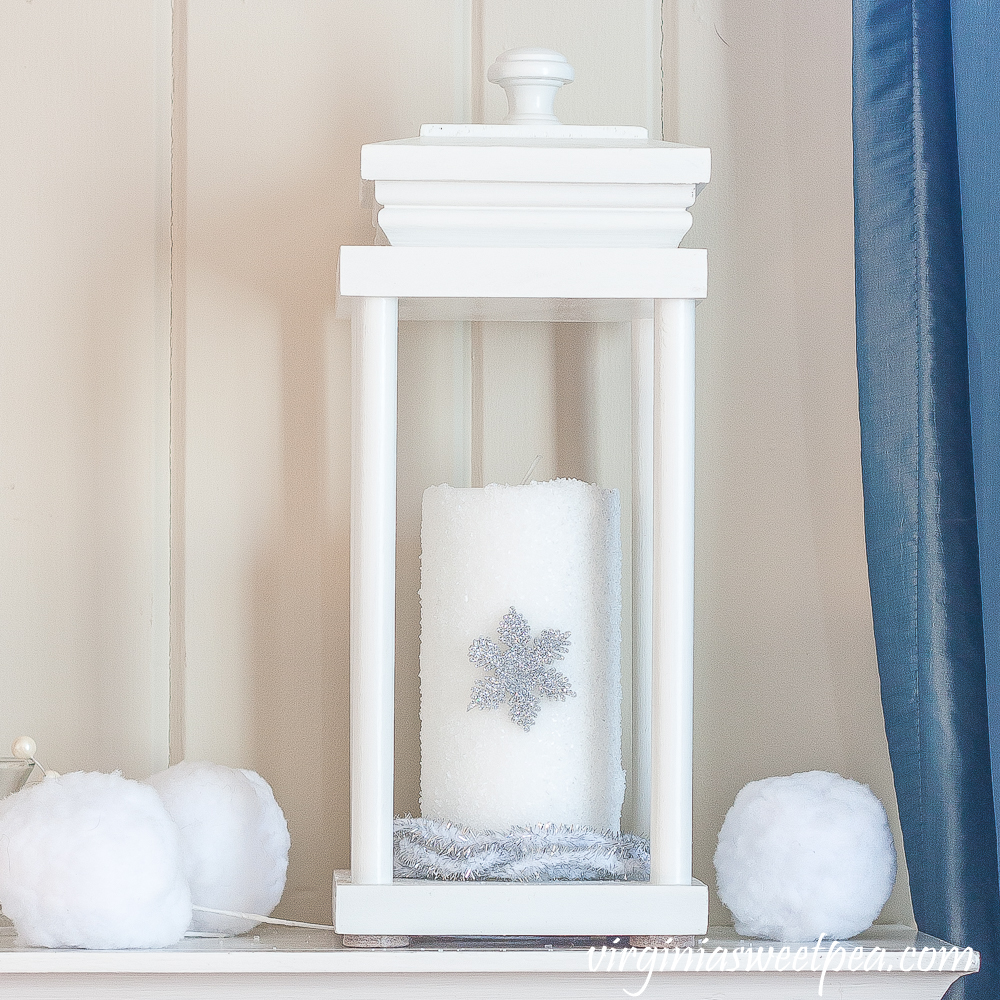 Lantern on a winter mantel with a candle covered in epsom salts with a snowflake surrounded by faux snowballs and faux berries.