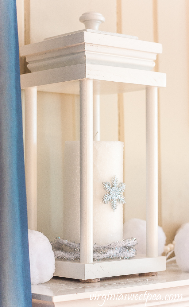 Lantern on a winter mantel with a candle covered in epsom salts with a snowflake surrounded by faux snowballs and faux berries.