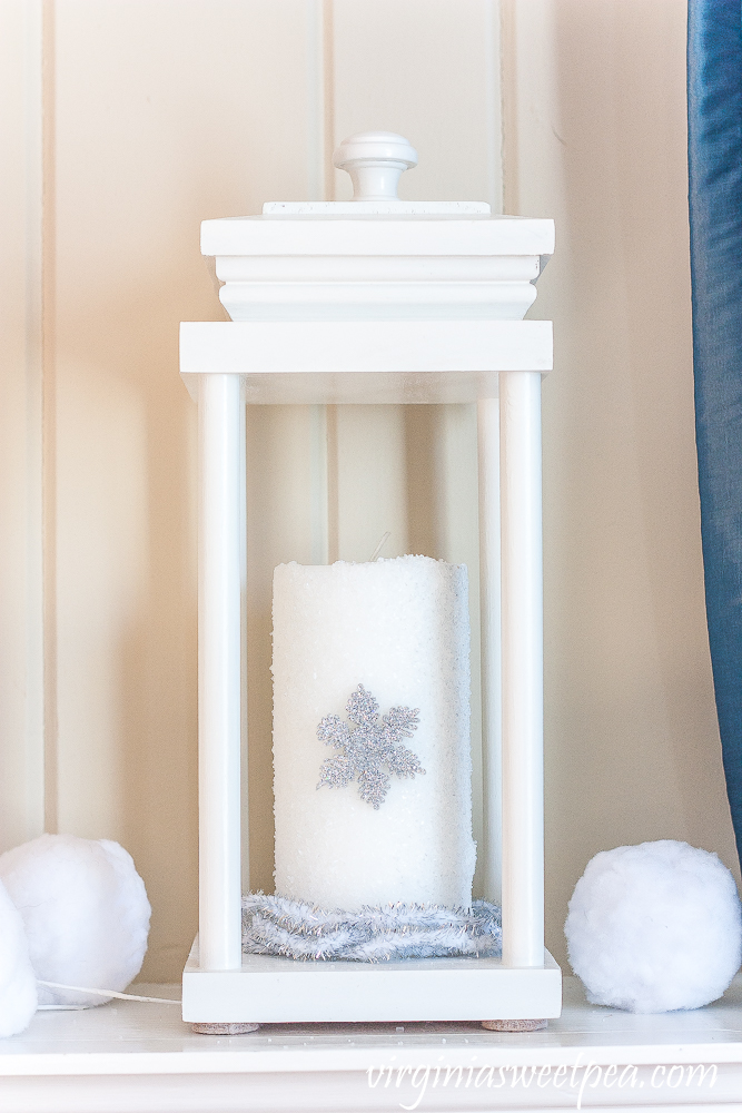 Lantern on a winter mantel with a candle covered in epsom salts with a snowflake surrounded by faux snowballs and faux berries.