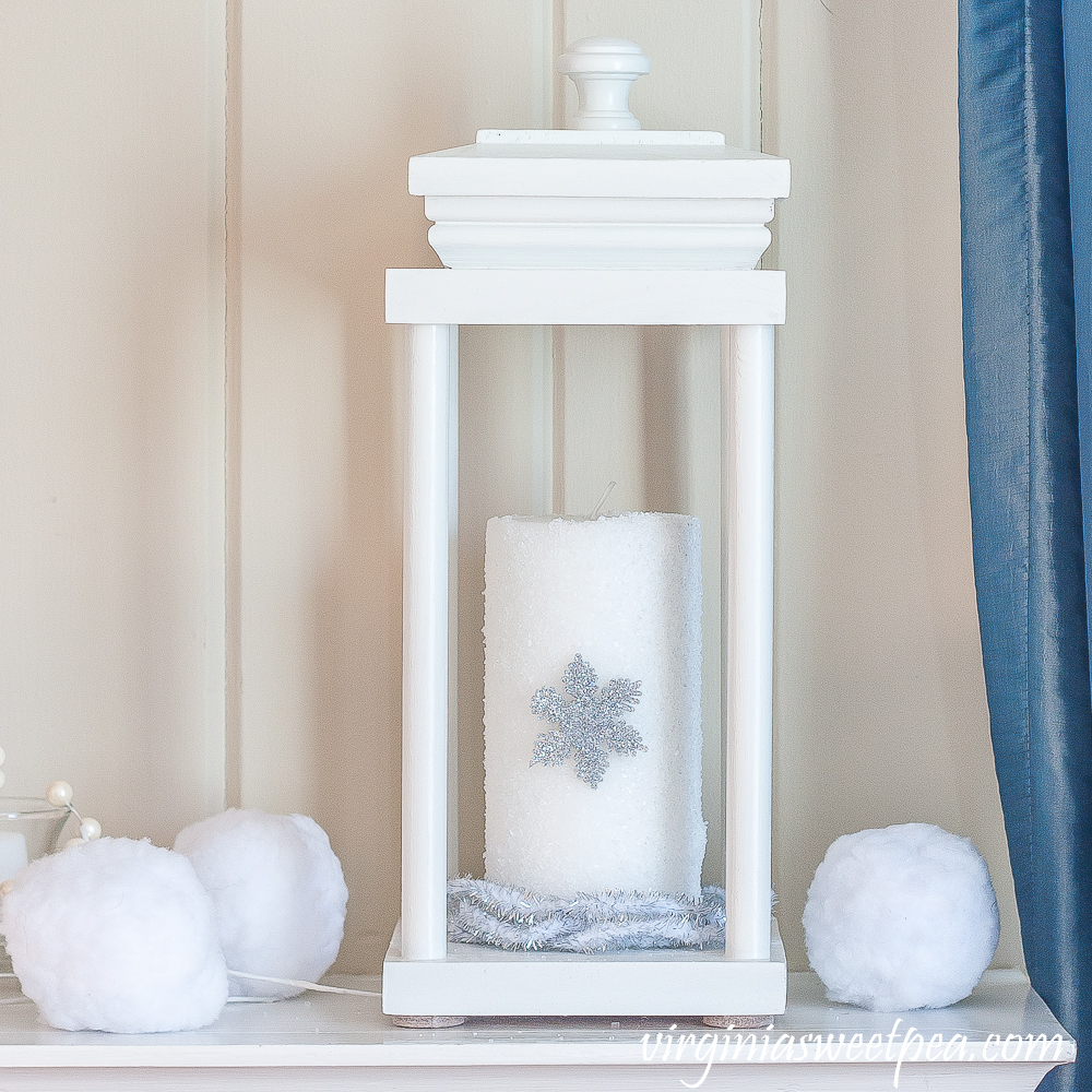 Lantern on a winter mantel with a candle covered in epsom salts with a snowflake surrounded by faux snowballs and faux berries.