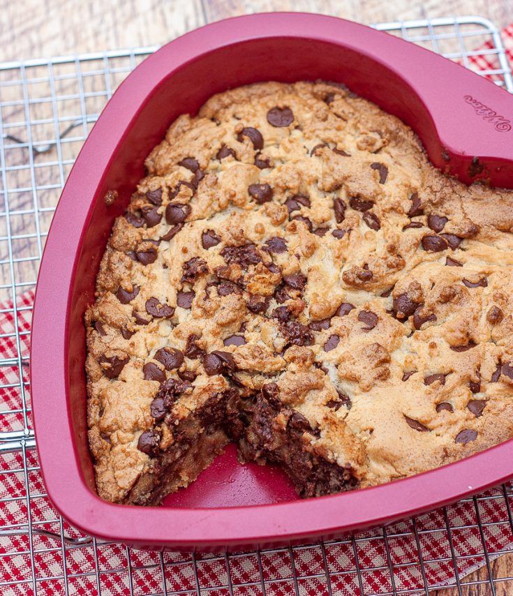 Chocolate chip cookie cake in a heart shaped pan on a wire rack with a red plaid napkin underneath