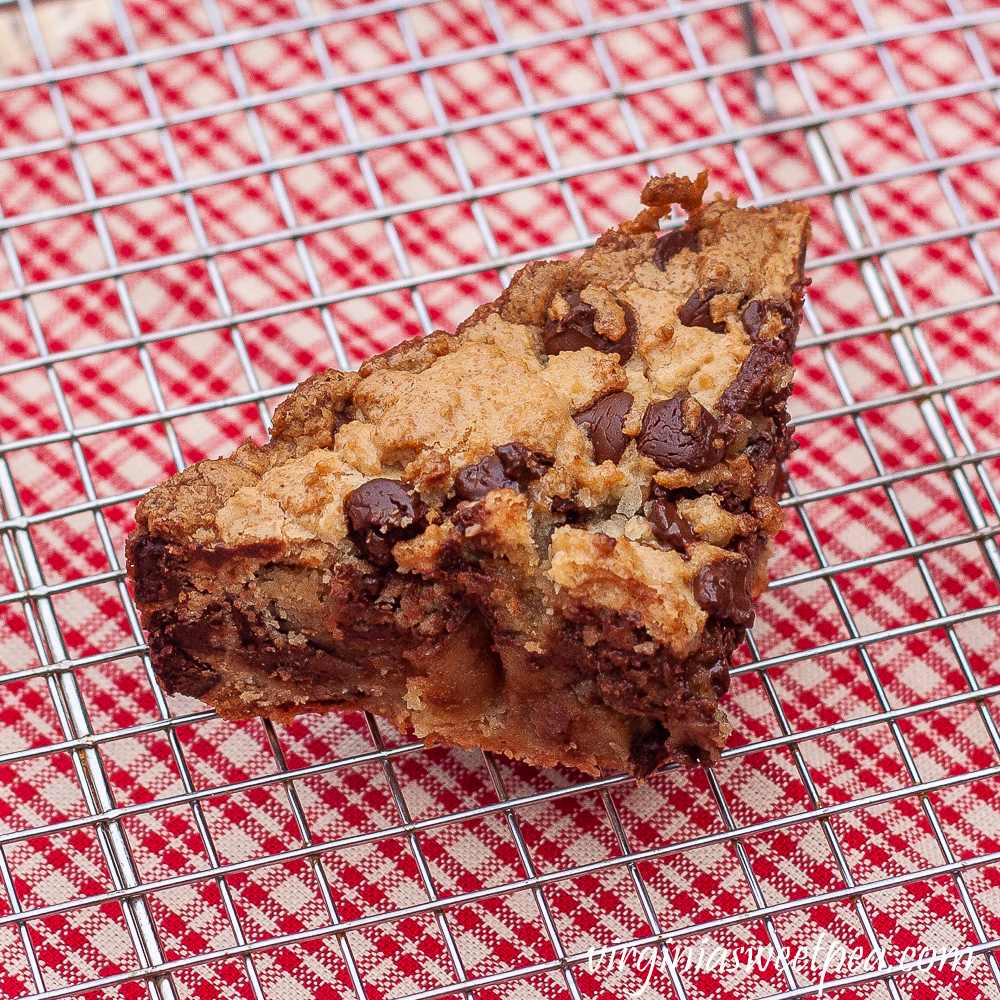 Piece of chocolate chip cookie cake on a wire rack with a red plaid napkin underneath