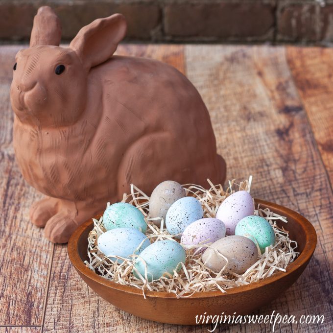 Speckled blue, green, and pink eggs in a wooden bowl filled with excelsior with a terracotta rabbit