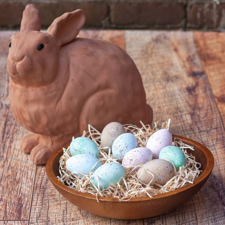 Speckled blue, green, and pink eggs in a wooden bowl filled with excelsior with a terracotta rabbit