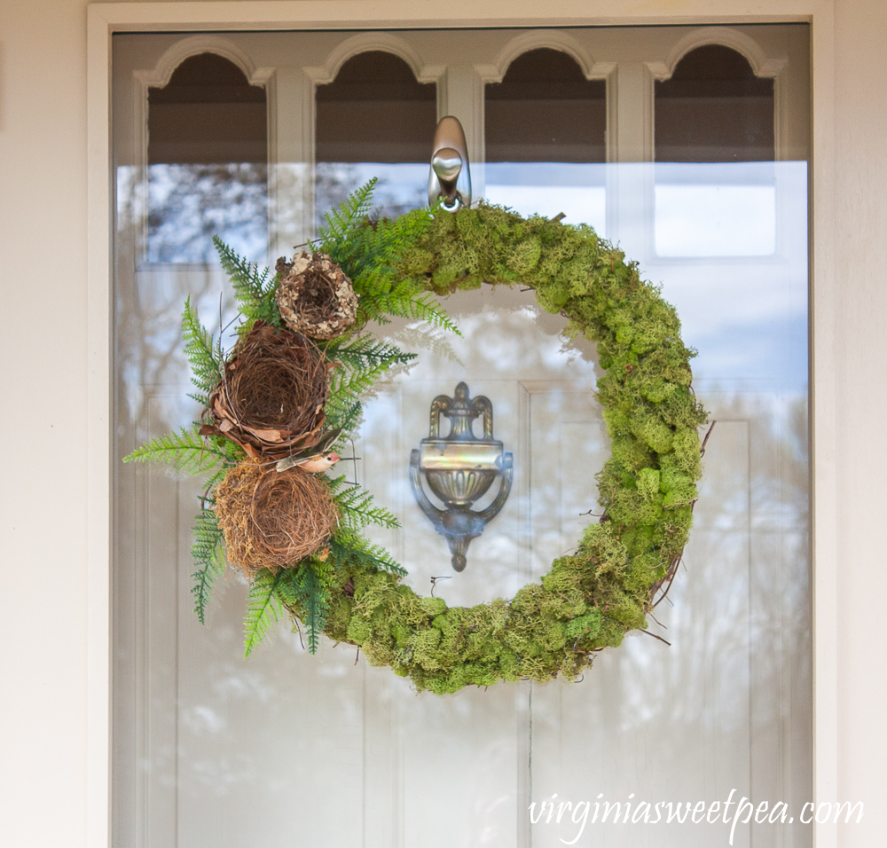 Wreath covered in moss with three nests, ferns, and a hummingbird on the side.