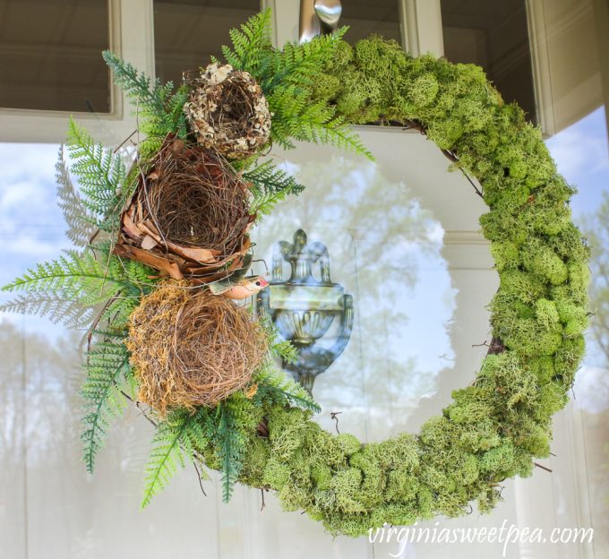 Wreath covered in moss with three nests, ferns, and a hummingbird on the side.