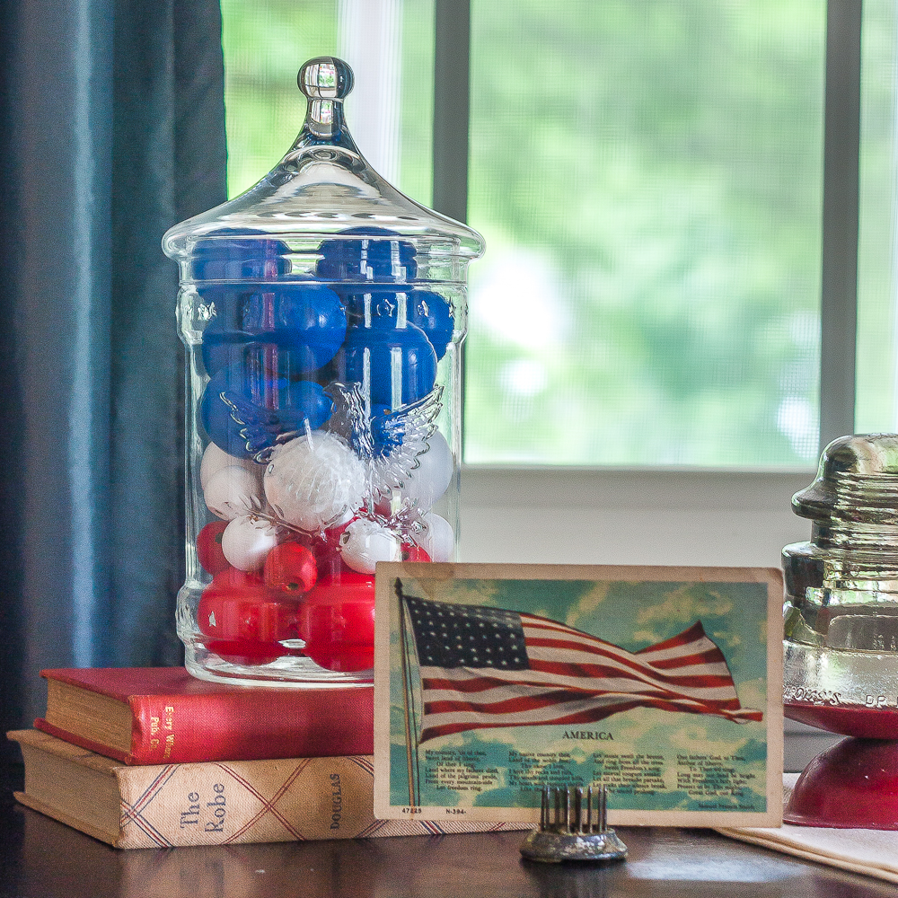 Patriotic Vignette with 1976 eagle jar with red, white, amd blue filler, American flag postcard, and glass insulator on a red candle stand on top of a vintage towel