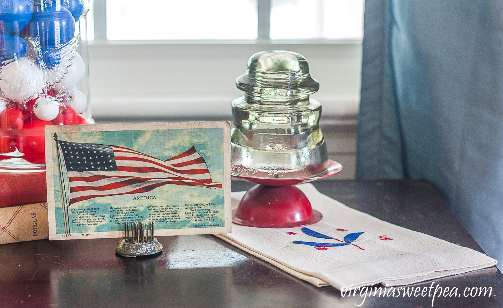 Patriotic Vignette with 1976 eagle jar with red, white, amd blue filler, American flag postcard, and glass insulator on a red candle stand on top of a vintage towel