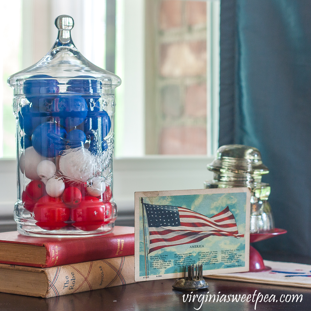 Patriotic Vignette with 1976 eagle jar with red, white, amd blue filler, American flag postcard, and glass insulator on a red candle stand on top of a vintage towel