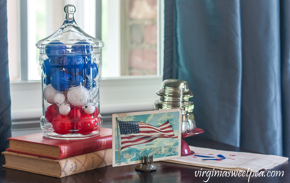 Patriotic Vignette with 1976 eagle jar with red, white, amd blue filler, American flag postcard, and glass insulator on a red candle stand on top of a vintage towel