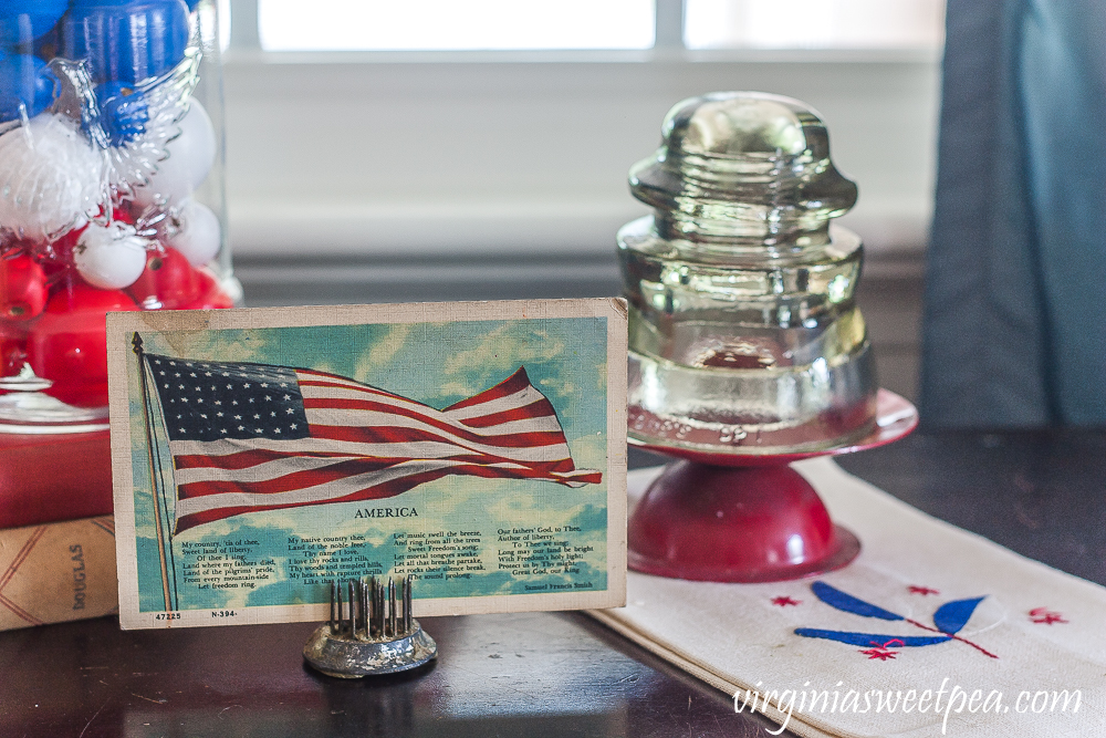 Patriotic Vignette with 1976 eagle jar with red, white, amd blue filler, American flag postcard, and glass insulator on a red candle stand on top of a vintage towel