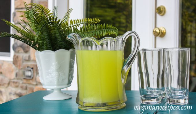 Lemonade ready to be served on an outdoor bar with glasses and a fern in a milk glass vase.