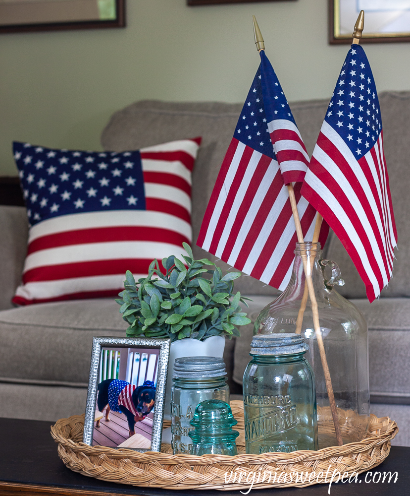 Patriotic Vignette with vintage Mason jars, vintage clear glass jug holding flags, and white milk glass compote with a faux plant and an American flag pillow in the background