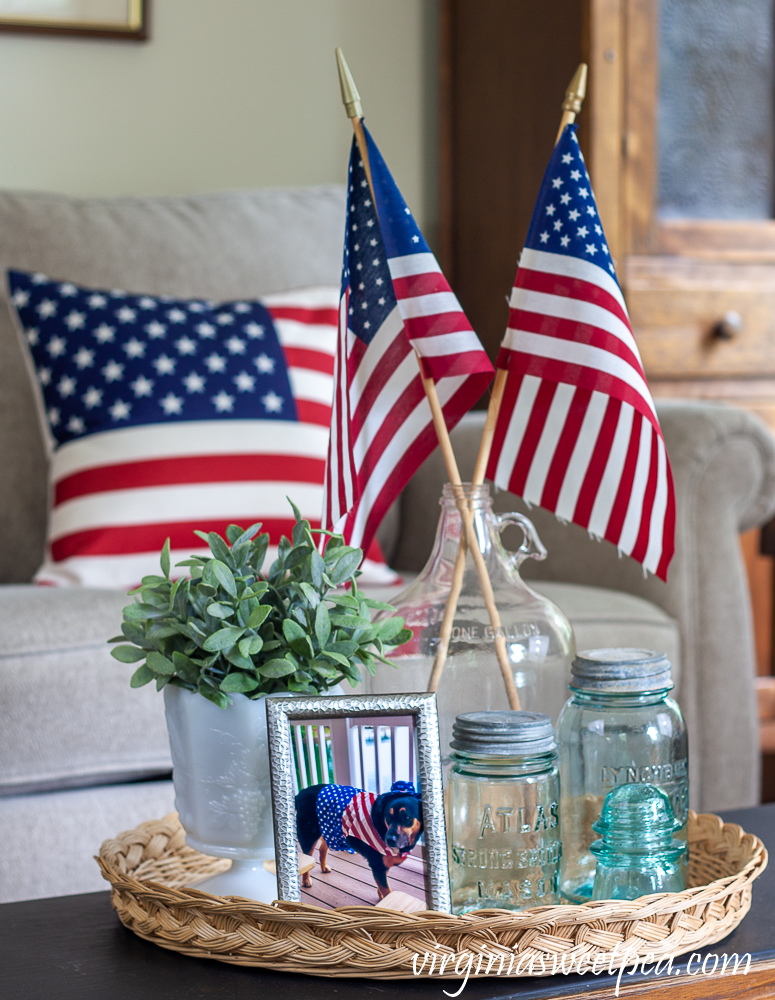 Patriotic Vignete with vintage Mason jars, vintage clear glass jug holding flags, and white milk glass compote with a faux plant and an American flag pillow in the background