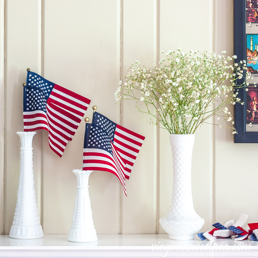 Milk glass vases with American flags and Baby's Breath on a mantel decorated patriotically.