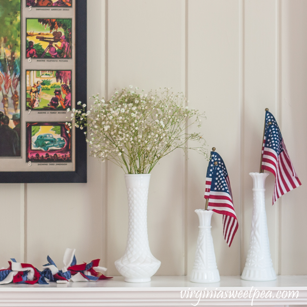 Milk glass vases with American flags and Baby's Breath on a mantel decorated patriotically.