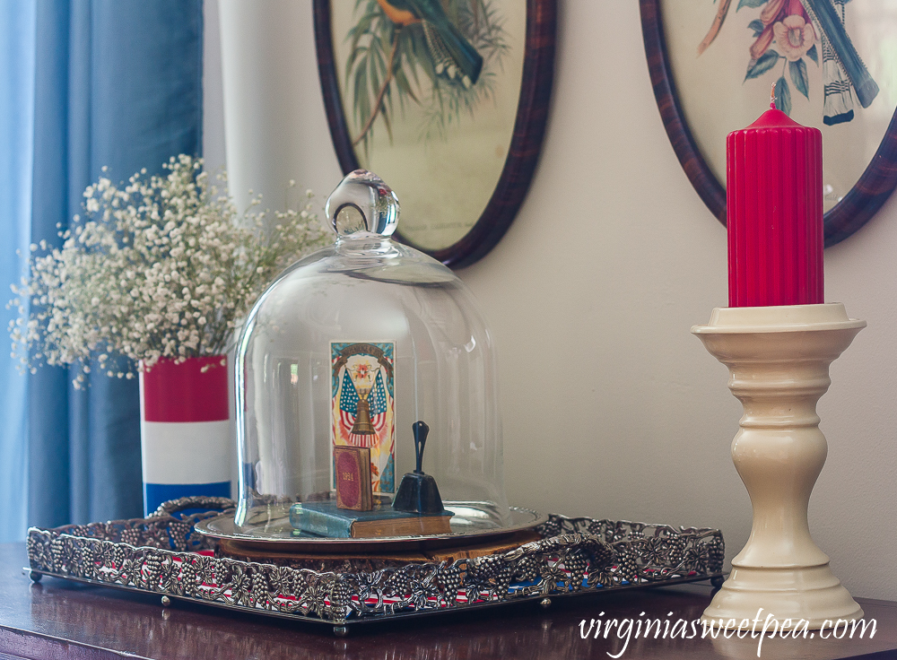 Patriotic Vignette with a red, white and blue striped vase with Baby's Breath and a cloche with a Liberty Bell postcard, 1924 mini book, a vintage bell, and a vintage bible with red candle on a cream colored candle holder.