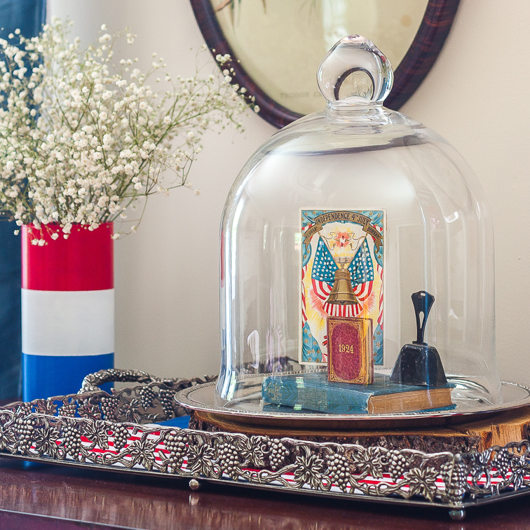 Patriotic Vignette with a red, white and blue striped vase with Baby's Breath and a cloche with a Liberty Bell postcard, 1924 mini book, a vintage bell, and a vintage bible.