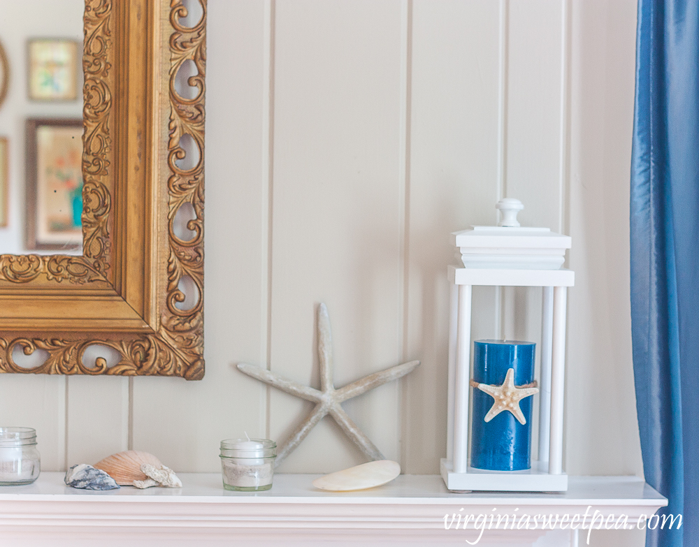 Coastal Themed summer mantel with a lantern holding a blue candle with a starfish tied to it, starfish, shells, coral, and small jars filled with sand and white votive candles.