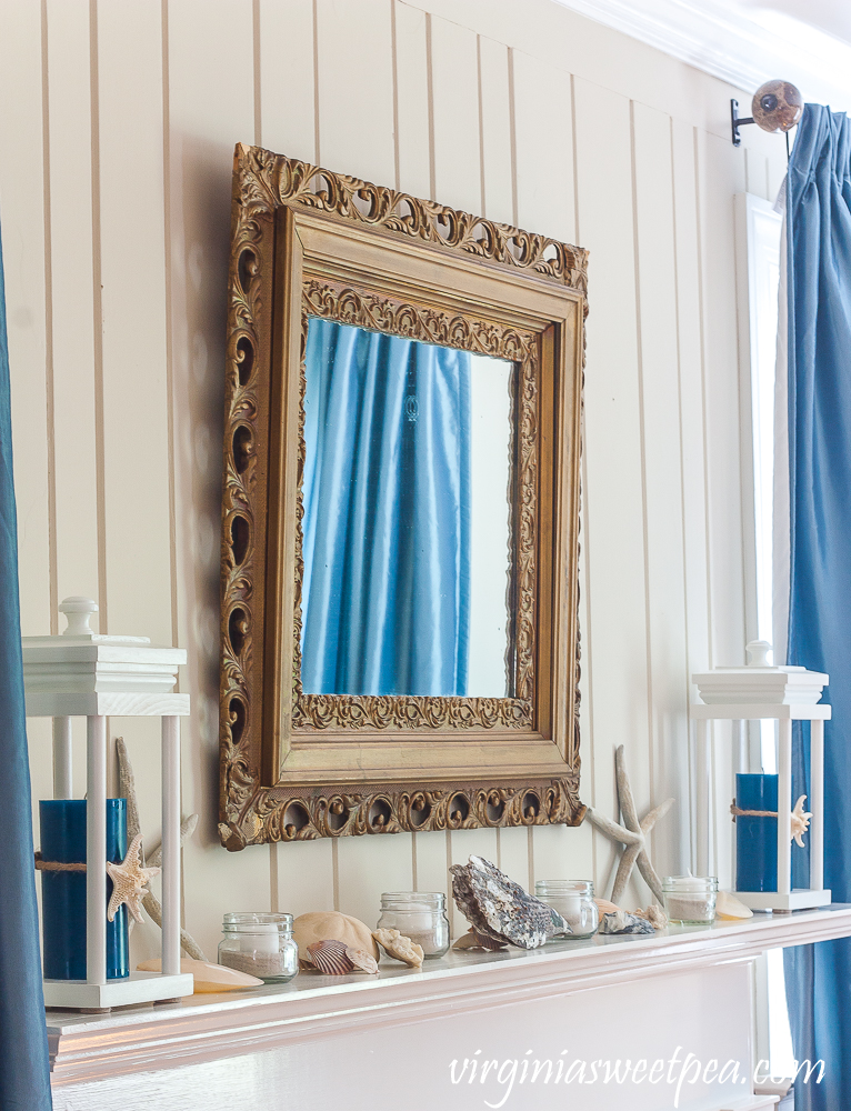 Coastal Themed summer mantel with a lantern holding a blue candle with a starfish tied to it, starfish, shells, coral, and small jars filled with sand and white votive candles.