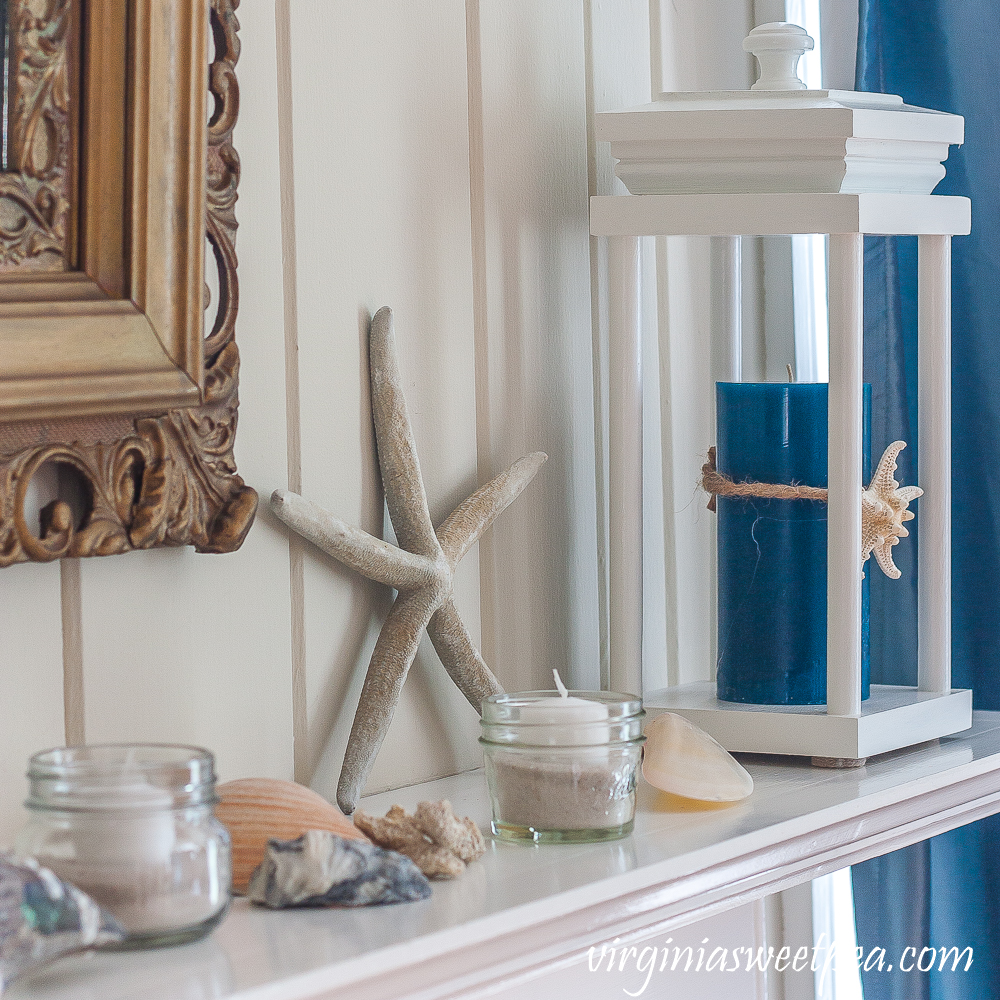 Coastal Themed summer mantel with a lantern holding a blue candle with a starfish tied to it, starfish, shells, coral, and small jars filled with sand and white votive candles.