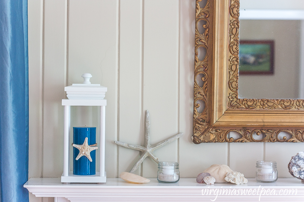 Coastal Themed summer mantel with a lantern holding a blue candle with a starfish tied to it, starfish, shells, coral, and small jars filled with sand and white votive candles.