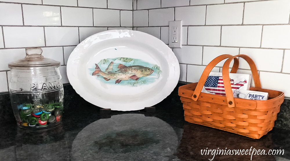 Longaberger basket on a kitchen counter with a Planter's Peanut jar filled with bottle caps and a vintage fish platter.